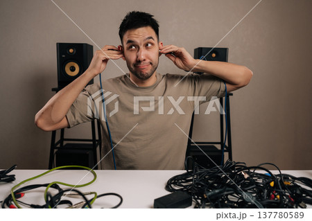 Man with an ironic expression struggling to manage numerous tangled audio cables on a table, holding one connection to each ear, surrounded by studio monitors and sound equipment. Man with an ironic expression struggling to manage numerous tangled audio cables on a table, holding one connection to each ear, surrounded by studio monitors and sound equipment. 137780589