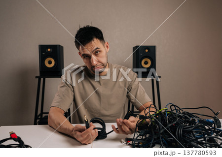 Portrait of frustrated man dealing with a massive pile of tangled electronic cables, facing a complex problem of wire management and technological clutter accumulation. Portrait of frustrated man dealing with a massive pile of tangled electronic cables, facing a complex problem of wire management and technological clutter accumulation. 137780593