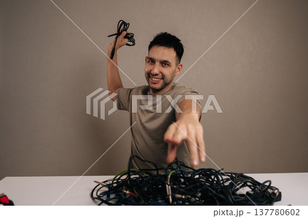 Vertical portrait of cheerful technician holding a large chaotic pile of tangled wires and cords, pointing finger directly forward, experiencing tech stress and cable management problems. Vertical portrait of cheerful technician holding a large chaotic pile of tangled wires and cords, pointing finger directly forward, experiencing tech stress and cable management problems. 137780602