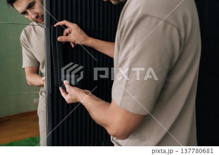 Contractor male installing a black acoustic foam panel, carefully fitting the soundproofing material on a wall surface to create an insulated environment. Contractor male installing a black acoustic foam panel, carefully fitting the soundproofing material on a wall surface to create an insulated environment. 137780681