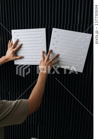 Vertical cropped shot of handyman positioning white acoustic insulation panels on a black soundproof foam wall, preparing a studio for optimal audio performance and effective sound dampening. Vertical cropped shot of handyman positioning white acoustic insulation panels on a black soundproof foam wall, preparing a studio for optimal audio performance and effective sound dampening. 137780694