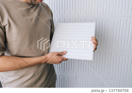 Man holding a square acoustic panel, preparing to install soundproofing material onto a textured white wall, focusing on noise reduction and enhancing room acoustics. 137780708