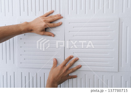 Close-up hand of handyman applying white soundproof acoustic panels onto a wall, improving home interior acoustics and soundproofing a room during renovation or construction. Close-up hand of handyman applying white soundproof acoustic panels onto a wall, improving home interior acoustics and soundproofing a room during renovation or construction. 137780715