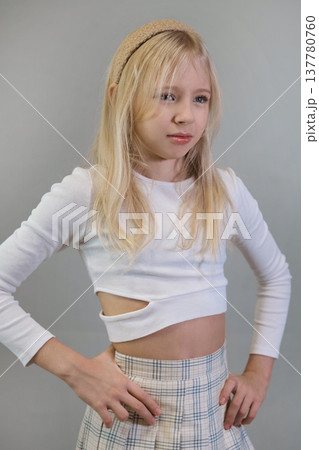 A young girl with blonde hair, wearing a white crop top and plaid skirt, poses confidently with her hands on her hips against a neutral background. This image highlights youthful fashion and a strong 137780760