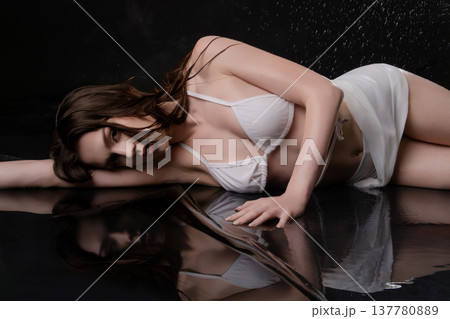 Sensual young woman in white bikini lying on a wet reflective black surface with water drops. 137780889