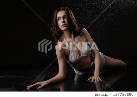 Dramatic studio portrait of a woman in a white bikini lying in water on a black background. 137780890