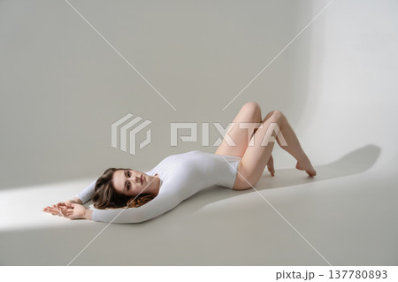 Elegant woman in a white long-sleeve bodysuit lying on a minimalist studio floor. 137780893