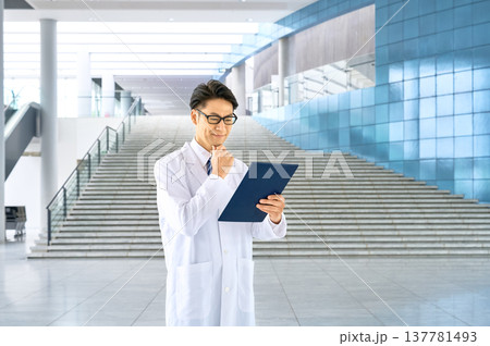 病院のロビーでカルテを確認して微笑む白衣の若い男性医師 137781493