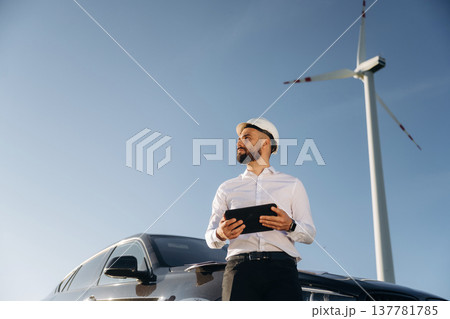 Leaning on the car, holding digital tablet. Engineer in field checking on wind turbine production Leaning on the car, holding digital tablet. Engineer in field checking on wind turbine production 137781785