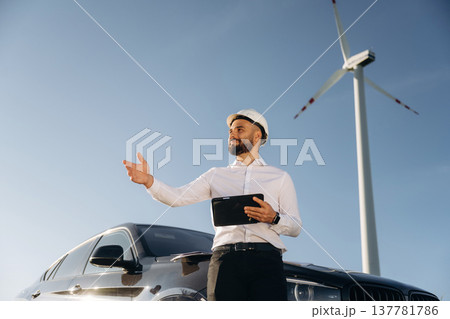 Leaning on the car, holding digital tablet. Engineer in field checking on wind turbine production Leaning on the car, holding digital tablet. Engineer in field checking on wind turbine production 137781786