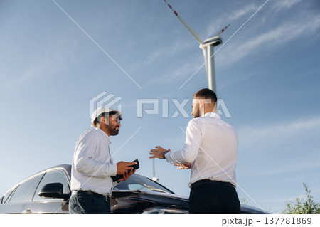 Black car, standing near it and talking. Two men engineers are on field, with wind turbines 137781869