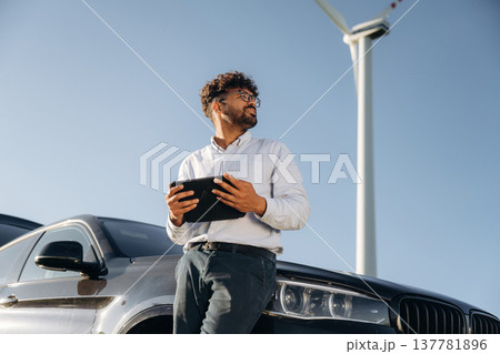 By the front of the car, with tablet. Male engineer in field checking on wind turbine production By the front of the car, with tablet. Male engineer in field checking on wind turbine production 137781896