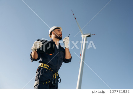 Speaking by walkie talkie. Engineer in field checking on wind turbine production Speaking by walkie talkie. Engineer in field checking on wind turbine production 137781922