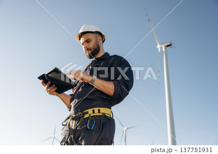 Professional uniform, hard hat and tablet. Engineer in field checking on wind turbine production Professional uniform, hard hat and tablet. Engineer in field checking on wind turbine production 137781931