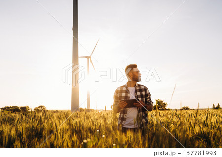 Sunny weather, front view. Guy engineer in wheat field against wind turbines Sunny weather, front view. Guy engineer in wheat field against wind turbines 137781993