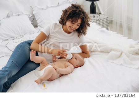 Giving food by using bottle. Young woman is with her young baby daughter indoors Giving food by using bottle. Young woman is with her young baby daughter indoors 137782164
