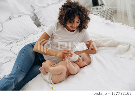 Giving food by using bottle. Young woman is with her young baby daughter indoors Giving food by using bottle. Young woman is with her young baby daughter indoors 137782165