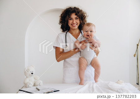 On the table and in hands, trying to stand up. Female doctor is with newborn baby indoors 137782203