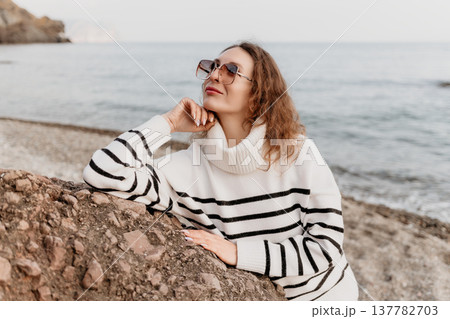 Woman beach sweater leaning on a rock looking at the sea during a serene day by the ocean with copy space Woman beach sweater leaning on a rock looking at the sea during a serene day by the ocean with copy space 137782703