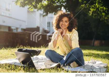 Looking forward, sitting on the blanket with laptop. Beautiful woman student is near the university outdoors Looking forward, sitting on the blanket with laptop. Beautiful woman student is near the university outdoors 137783567