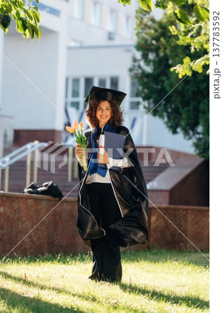 Green tree and university building behind. Cheerful graduate student in a gown is standing outdoors Green tree and university building behind. Cheerful graduate student in a gown is standing outdoors 137783592