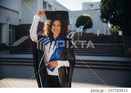 Feeling good, holding diploma. Cheerful graduate student in a gown is standing outdoors 137783615