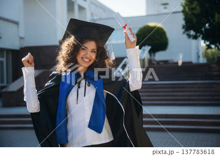 Diploma in hand. Cheerful graduate student in a gown is standing outdoors 137783618