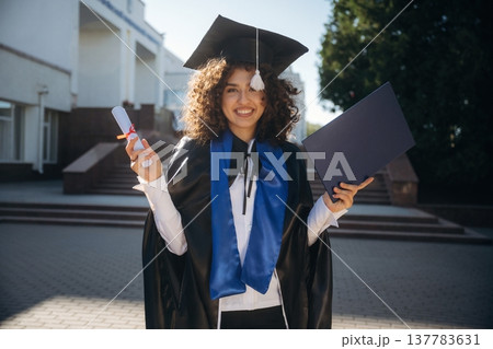 Feeling good, holding diploma. Cheerful graduate student in a gown is standing outdoors 137783631