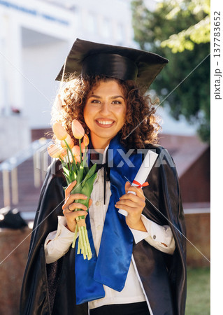 Flowers in hand. Cheerful graduate student in a gown is standing outdoors 137783652