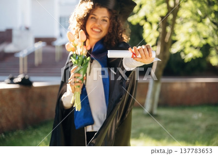 Flowers in hand. Cheerful graduate student in a gown is standing outdoors Flowers in hand. Cheerful graduate student in a gown is standing outdoors 137783653