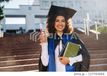 Standing against university building. Cheerful graduate student in a gown is standing outdoors Standing against university building. Cheerful graduate student in a gown is standing outdoors 137783669