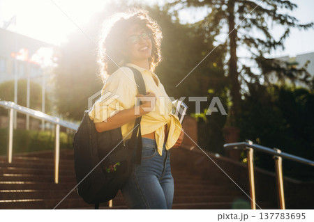 Under bright sunlight, smiling. Beautiful woman student is near the university outdoors Under bright sunlight, smiling. Beautiful woman student is near the university outdoors 137783695