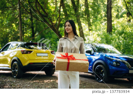 Present box in hands, standing. Young woman is in the forest with two cars Present box in hands, standing. Young woman is in the forest with two cars 137783934