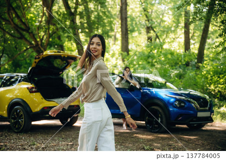 Posing and standing. Two women are near yellow and blue cars in the forest Posing and standing. Two women are near yellow and blue cars in the forest 137784005
