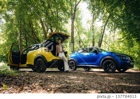 Vacation conception. Holding drink in hands. Young woman is in the forest with two cars Vacation conception. Holding drink in hands. Young woman is in the forest with two cars 137784013