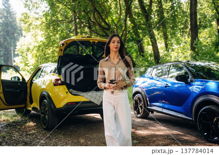 Vacation conception. Holding drink in hands. Young woman is in the forest with two cars Vacation conception. Holding drink in hands. Young woman is in the forest with two cars 137784014