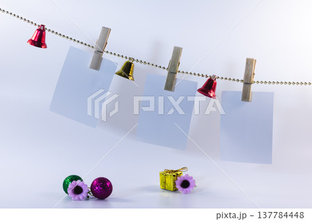 Blank photo cards hanging on string with wooden clips and colorful bells. Festive decoration with glitter balls, small gift and flowers on white background with copy space. Blank photo cards hanging on string with wooden clips and colorful bells. Festive decoration with glitter balls, small gift and flowers on white background with copy space. 137784448