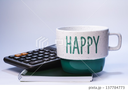 Ceramic coffee mug with Happy text placed beside calculator on spiral notebook isolated on white background. Business finance, accounting and productive workplace concept. Ceramic coffee mug with Happy text placed beside calculator on spiral notebook isolated on white background. Business finance, accounting and productive workplace concept. 137784773