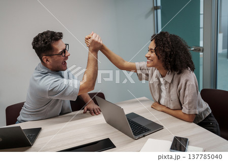 Friendly Coworkers Engage In Lighthearted Arm Wrestling During Casual Meeting Break With Laptops Nearby 137785040