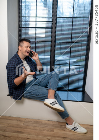 Laughing man sitting by window talking on smartphone in contemporary office 137785458