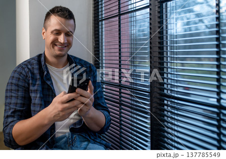 Content man using smartphone standing near window in modern office interior Content man using smartphone standing near window in modern office interior 137785549