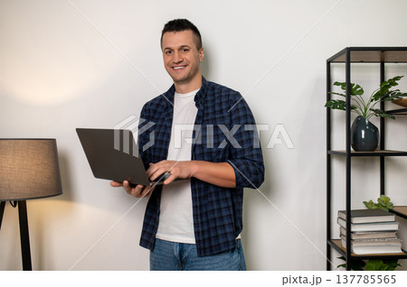 Smiling man holding laptop in home setting with shelf and lamp Smiling man holding laptop in home setting with shelf and lamp 137785565