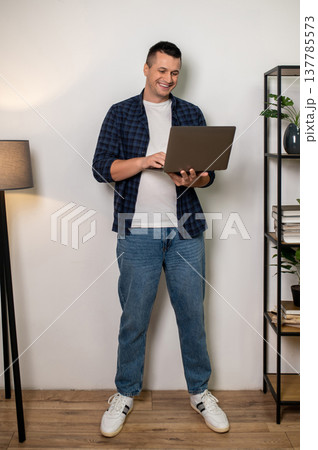 Cheerful man holding laptop standing in home interior with shelf and lamp Cheerful man holding laptop standing in home interior with shelf and lamp 137785573