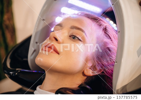Pink or velvet color. Woman is in a beauty salon with LED hair growth helmet Pink or velvet color. Woman is in a beauty salon with LED hair growth helmet 137785575