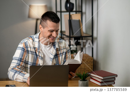Cheerful man reading book and working on laptop in modern home workspace 137785589