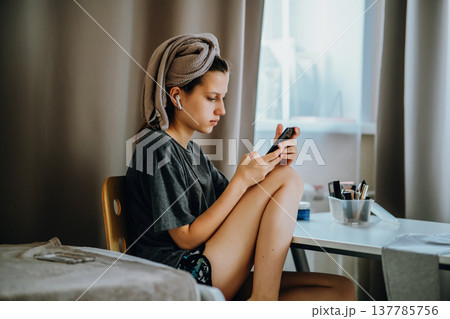 Teen girl sitting at home by the window with a smartphone, earphones and a bath towel on her head. Natural home lifestyle scene about messaging, music, privacy and everyday life. Teen girl sitting at home by the window with a smartphone, earphones and a bath towel on her head. Natural home lifestyle scene about messaging, music, privacy and everyday life. 137785756