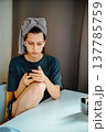 Teen girl sitting at a table at home and looking at her phone. The towel on her head and earphones create a story about music, communication, study and quiet daily life. 137785759