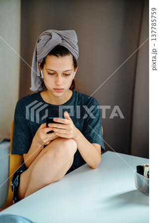 Teen girl sitting at a table at home and looking at her phone. The towel on her head and earphones create a story about music, communication, study and quiet daily life. Teen girl sitting at a table at home and looking at her phone. The towel on her head and earphones create a story about music, communication, study and quiet daily life. 137785759