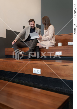 Two coworkers reviewing notes and discussing project on wooden benches in a neutral office setting. Review, notes, project, office, teamwork. Two coworkers reviewing notes and discussing project on wooden benches in a neutral office setting. Review, notes, project, office, teamwork. 137785783