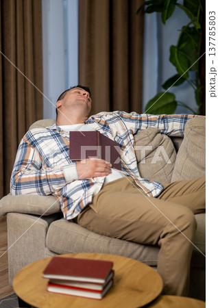 Tired man sleeping with book on sofa in modern living room interior Tired man sleeping with book on sofa in modern living room interior 137785803